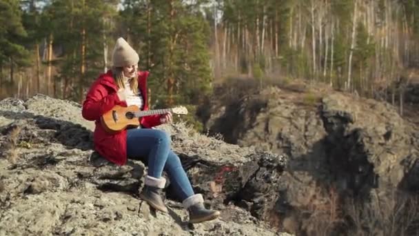 Biała kobieta siedzi na skale i gra na ukulele. Jej blond włosy wieje wiatr. Gra na strunach małej gitary. Jesień, żółte liście na drzewach. Zainteresowania, struny, muzyka — Wideo stockowe