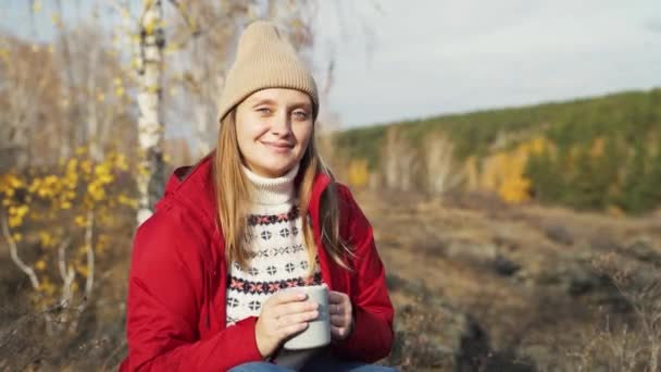 Caucasian woman is sitting alone in autumn forest, tasting the aroma of tea from gray mug and enjoying nature. She smiles. She is wearing red raincoat, beige hat and white sweater. Autumn, tea time — Stock Video