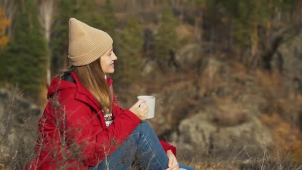 Mujer caucásica está sentado solo en un bosque de otoño, beber té caliente de taza gris y disfrutar de la naturaleza. El viento sopla suavemente su cabello rubio y ella sonríe. Senderismo, otoño, hora del té — Vídeos de Stock