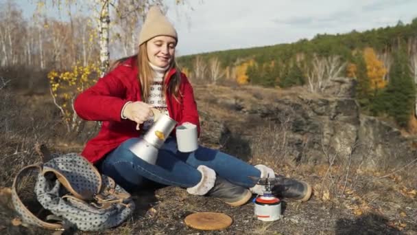 Kobieta siedzi sama w jesiennym lesie, wlewając świeżą gorącą herbatę do szarego kubka i ciesząc się naturą. Wiatr delikatnie rozwija liście na drzewach i uśmiecha się. Piesze wycieczki, jesień, czas na herbatę — Wideo stockowe