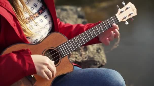 Fehér nő ül a sziklán és ukulelén játszik. A szőke haját fújja a szél. Kis gitárhúrokon játszik. Ősz, sárga levelek a fákon. Hobbi, húr, zene — Stock videók