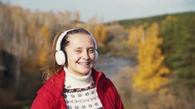 Beyaz kulaklıklarla tepelerde dans eden beyaz bir kadın. Diş telleri dişlerde görünür. Kırmızı ceket ve beyaz kazak giyiyor, güneşe bakıyor. Doğa, sonbahar, teknoloji