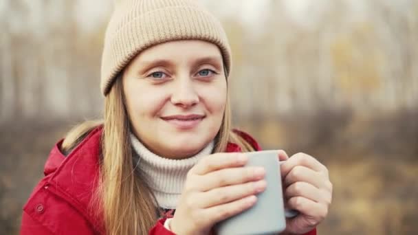 女性は秋の森の中に座って、灰色のマグカップから熱いお茶を飲んで自然を楽しんでいます。風はブロンドの髪を優しく吹く。彼女は赤いレインコート、ベージュの帽子をかぶっている。ハイキング、秋、ティータイム — ストック動画