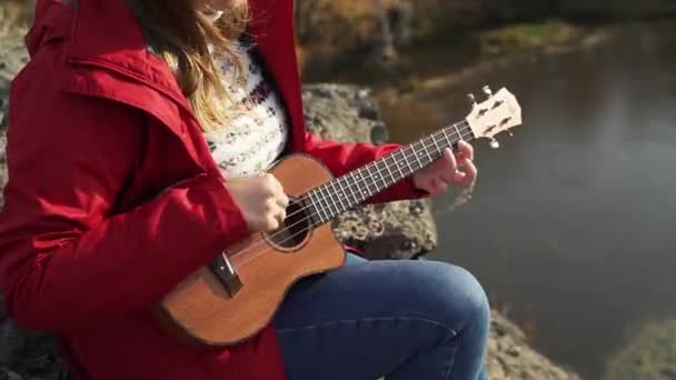 Mulher branca senta-se na rocha e toca ukulele. Seu cabelo loiro é soprado pelo vento. Ela toca as cordas de uma guitarra pequena. Outono, folhas amarelas nas árvores. Passatempos, cordas, música — Vídeo de Stock