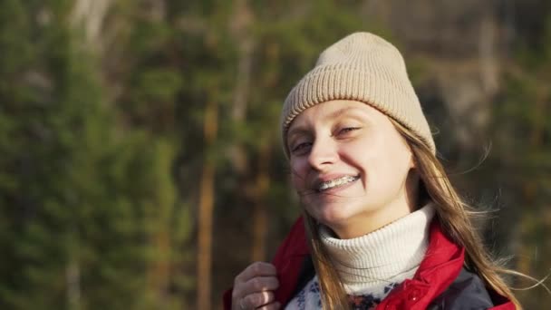 Biała kobieta w uśmiechach i aparacie na zębach. Mruży oczy na słońcu i śmieje się. Wiatr rozwija włosy i błyszczą w świetle. Jesienna wędrówka, natura, żółte drzewa, rzeka — Wideo stockowe