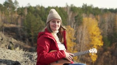 Kadın kayanın üzerine oturur ve ukulele tutar. Sarı saçları rüzgârla savruluyor. Güneş parlıyor ve o gülümsüyor. Sonbahar, ağaçlardaki sarı yapraklar. Hobiler