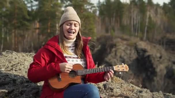 Woman sits on rock and plays the ukulele. Her blonde hair is blown by the wind. She portrays rock star and actively strikes the strings and sings. Autumn, yellow leaves on trees. Hobbies — Stock Video