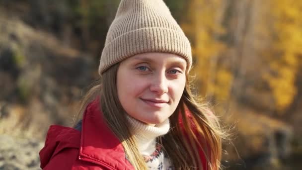 Mujer caucásica con ojos azules en sombrero beige sonríe románticamente. Está entrecerrando los ojos al sol. El viento desarrolla el cabello y brillan en la luz. Otoño, naturaleza, árboles amarillos — Vídeos de Stock