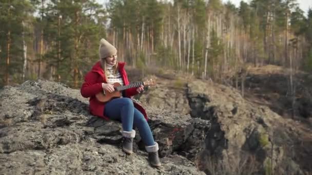 Mulher branca senta-se na rocha e toca ukulele. Seu cabelo loiro é soprado pelo vento. Ela toca as cordas de uma guitarra pequena. Outono, folhas amarelas nas árvores. Passatempos, cordas, música — Vídeo de Stock