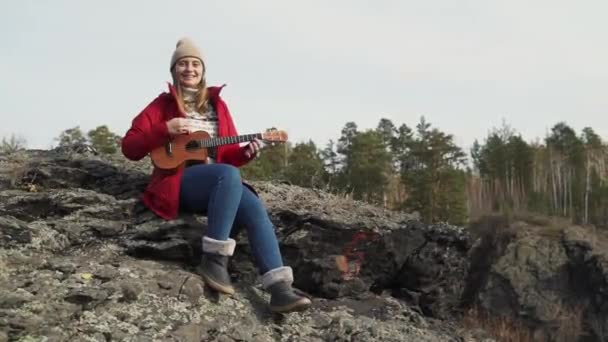 Kaukasierin sitzt auf Felsen und spielt Ukulele. Ihre blonden Haare werden vom Wind verweht. Sie spielt die Saiten einer kleinen Gitarre. Herbst, gelbe Blätter an Bäumen. Hobbys, Saiten, Musik — Stockvideo