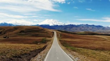 Kafkasya sırtı ve Elbrus Dağı manzaralı harika bir yol manzarası. Otoyol yeşil çayır boyunca uzanıyor. Hafif bulutlar Elbrus 'un beyaz karlı tepelerini ve mavi gökyüzünü kaplıyor.