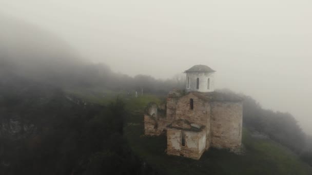 Aerial view of ancient Christian Sentinel Church, Caucasus. It is located on spur of mountain range. Thick fog covers white stone walls of church and greenery around. Roof is covered in moss — Stock Video
