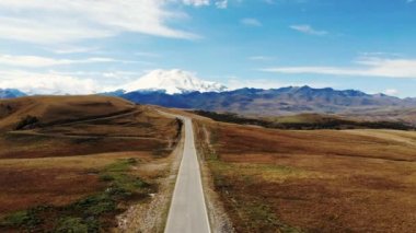 Kafkasya sırtı ve Elbrus Dağı manzaralı harika bir yol manzarası. Otoyol yeşil çayır boyunca uzanıyor. Hafif bulutlar Elbrus 'un beyaz karlı tepelerini ve mavi gökyüzünü kaplıyor.
