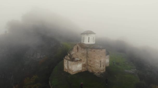 Повітряний вид старовинної християнської церкви Сентінел, Кавказ. Розташований на хребті. Густий туман покриває білі кам'яні стіни церкви і зелень навколо. Дах покритий мохом. — стокове відео
