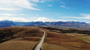 Kafkasya sırtı ve Elbrus Dağı manzaralı harika bir yol manzarası. Otoyol yeşil çayır boyunca uzanıyor. Hafif bulutlar Elbrus 'un beyaz karlı tepelerini ve mavi gökyüzünü kaplıyor.