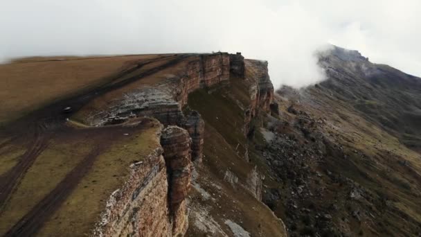 Повітряний вид кам'яного амфітеатру в товстих хмарах, Bermamyt plateau, Caucasus. Летючи над величною скелею, вздовж якої пролягає дорога. Туман рухається вгору по крутому схилу. Ельбрус — стокове відео