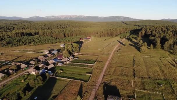 Luchtfoto van het Russische dorp in de zomer. Vliegen over houten huizen, dicht groen en velden in kleine Russische nederzetting. Zon verlicht de bergen en het dichte groene bos in de verte — Stockvideo