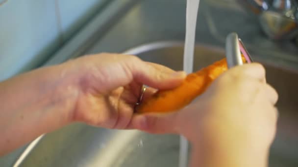 La donna stacca leggermente le giovani carote mature con pelapatate sopra il lavandino d'argento vicino all'acqua che scorre in cucina. Il dispositivo in metallo rimuove lo strato sottile. Note di alimentazione, verdure — Video Stock
