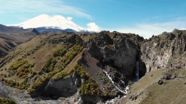 Luftaufnahme des Bergwasserfalls und des Elbrus. Blick auf den schneeweißen wolkenlosen Gipfel des Elbrus. Stürmischer Fluss fließt vom Gipfel des Berges in Schlucht und bricht am Boden auf — Stockvideo