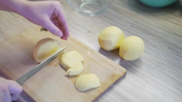 La mujer está rebanando finamente papas frescas jóvenes en la cocina con un cuchillo afilado en una tabla de madera para cocinar. Pequeñas piezas caen pieza por pieza en el tablero. Alimentación saludable, verduras, cocinar en casa — Vídeo de stock