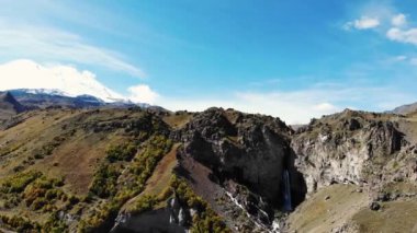Dağ şelalesi ve Elbrus Dağı 'nın havadan görünüşü. Elbrus Dağı 'nın karlı, bulutsuz zirvesinin manzarası. Fırtınalı nehir dağın tepesinden vadiye akıyor ve yerden kırılıyor.