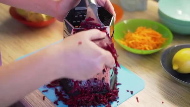 Mujer muele remolacha borgoña en rallador de metal plateado en la cocina en tablero azul para cocinar. En el fondo hay platos coloridos con verduras. Comida saludable, verduras, cocinar en casa — Vídeos de Stock