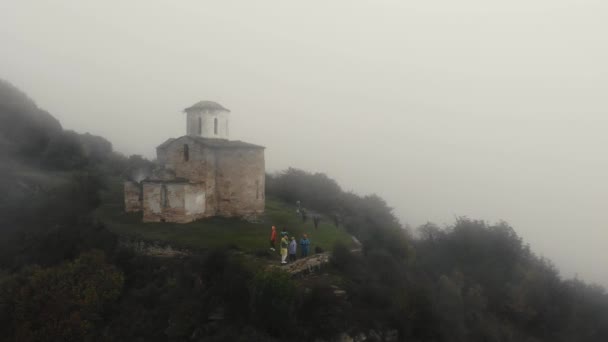 Letecký pohled na starověkou křesťanskou Sentinelovou církev na Kavkaze. Nachází se na úpatí pohoří. Bílé kamenné zdi kostela a okolí pokrývá hustá mlha. Střecha je pokrytá mechem — Stock video