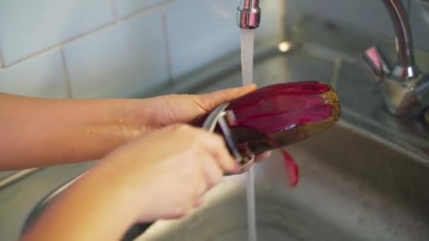 Kvinnan tar lätt bort skalet från mogna unga rödbetor med grönsaksskalare över silvervasken nära det rinnande vattnet i köket. Metallanordning avlägsnar tunt lager. Hälsosam kost, vegetarism — Stockvideo