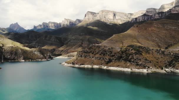 Luchtfoto van het azuurblauwe bergmeer Gizhgit, Kaukasus. Blauw water schijnt in het zonlicht. Hoge majestueuze bergketen is zichtbaar. Scherpe pieken van pieken gloeien tegen de bewolkte lucht. Drone videoshooting — Stockvideo