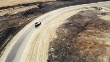 İspatlama alanı boyunca giden kamyonetin hava görüntüsü. Sürücü yeni yolun kalitesini kontrol ediyor. Kamyonlar ve işçiler yakınlarda duruyor. Yol işaretli asfalt. Yol yapımı