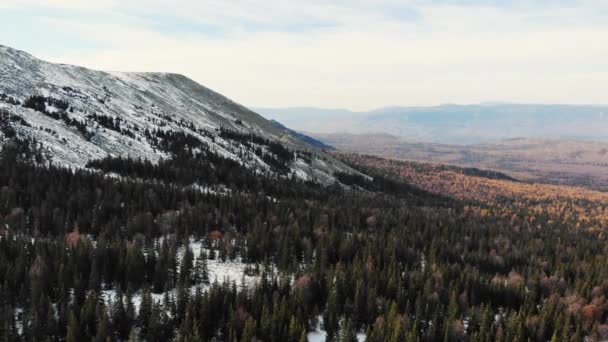 Vista aérea das encostas das montanhas cobertas com floresta de abeto e a primeira neve, o Ural do Sul. A cordilheira pode ser vista na névoa de luz à distância. O céu azul está coberto de nuvens — Vídeo de Stock