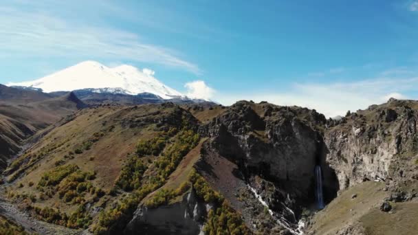 Luftaufnahme des Bergwasserfalls und des Elbrus. Blick auf den schneeweißen wolkenlosen Gipfel des Elbrus. Stürmischer Fluss fließt vom Gipfel des Berges in Schlucht und bricht am Boden auf — Stockvideo