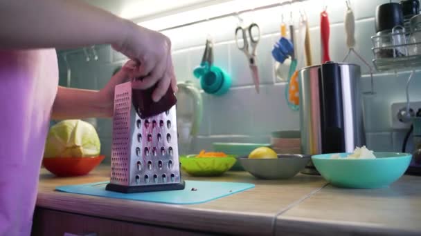 Женщина шлифует бордовую свеклу на серебряной металлической терке на кухне на голубой доске для приготовления пищи. На заднем плане - красочные тарелки с овощами. Здоровое питание, овощи, приготовление пищи на дому — стоковое видео