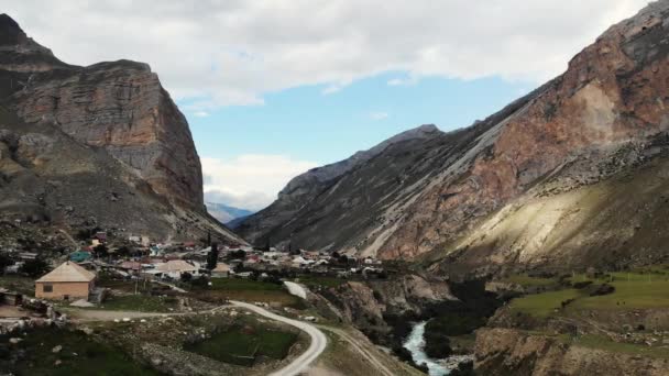 Повітряний вид на село, розташоване між горами Кавказу. Розташоване біля річки. Вершини гір дуже високі і суворі. Смуги покриті камінням і невеликою зеленою рослинністю. — стокове відео