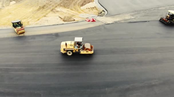 Vista aérea dos rolos de estrada amarelos que colocam o asfalto do terreno de teste para carros. Carros rolar suavemente asfalto preto ao longo dos caminhos. Outro equipamento de construção nas proximidades. Construção de estradas — Vídeo de Stock