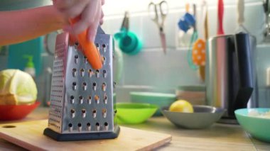 Kadın yemek pişirmek için mutfaktaki gümüş metal rende turuncu havuçları öğütüyor. Arka planda sebzeli renkli tabaklar var. Sağlıklı yiyecekler, sebzeler, evde yemek pişirmek.