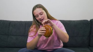 Mavi gözlü ve pembe tişörtlü tombul genç bir kadın gri koltukta oturuyor ve büyük bir zevkle koca bir hamburger yiyor. Kolesterol, abur cubur, alkol, sağlıksızlık, oburluk yağı