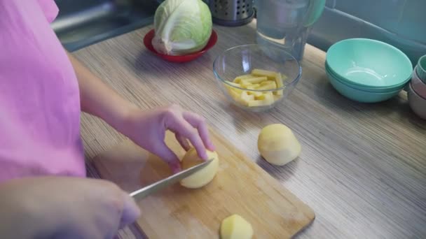 女性は、調理のための木製ボード上の鋭いナイフで台所で若い新鮮なジャガイモを細かくスライスしています。小片は板の上に一枚ずつ落ちる。健康的な食事、野菜、家庭での料理 — ストック動画