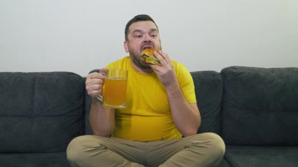 Пухнастий молодий чоловік сидить на сірому дивані і їсть великий жирний бургер і п'є пиво з великого скляного горнятка. Він кусає булочку у великі шматки. Холестерин, нездорове харчування, алкоголь, нездорове здоров'я, пухнастий жир — стокове відео