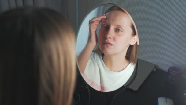 Die junge Frau sieht sich selbst im Spiegel an, untersucht die Haut ihres Gesichts und regt sich auf. Es gibt Rötungen und Pickel im Gesicht. Das Mädchen ist aufgebracht. Gesundheit, Kosmetologie — Stockvideo