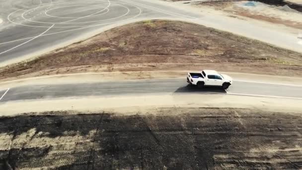 Widok z lotu ptaka białego pickupa jadącego ze wzgórza na poligonie doświadczalnym. Kierowca samochodu sprawdza jakość nowej drogi. Czarny asfalt ze znakami drogowymi. Budowa dróg, poligon doświadczalny — Wideo stockowe