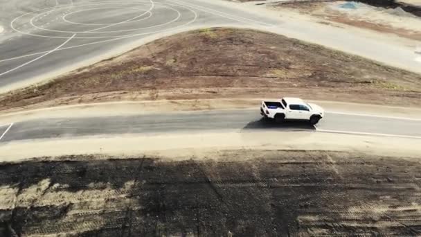 証明された地面で丘を運転白いピックアップトラックの空中ビュー。車の運転手は新しい道路の品質をチェックします。道路標識付き黒アスファルト。道路建設、試験場 — ストック動画