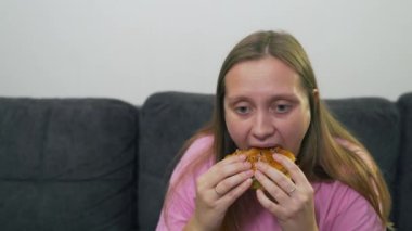 Şişman, mavi gözlü, depresyondaki pembe tişörtlü beyaz bir kadın gri kanepede oturuyor ve koca bir hamburger yiyor. Yüzü yorgun ve üzgün. Kolesterol, sağlıksız diyet, oburluk yağı.