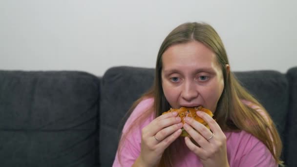 Πλούσια νεαρή καυκάσιος γυναίκα με μπλε μάτια και ένα ροζ T-shirt σε κατάθλιψη κάθεται σε γκρι καναπέ και τρώει μεγάλο λίπος burger. Το πρόσωπό της είναι κουρασμένο και λυπημένο. Χοληστερόλη, ανθυγιεινή διατροφή, λίπος λαιμαργίας — Αρχείο Βίντεο