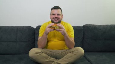 Şişman, kahverengi gözlü genç adam gri koltukta oturuyor ve büyük, şişman bir hamburger yiyor. Büyük parçalar halinde topuzu ısırıyor. Susuzluk ve açlık. Kolesterol, sağlıksız beslenme, sağlıksız beslenme, oburluk yağı.