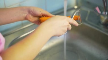 Kadın, taze taze havuçları gümüş bir lavabo üzerinde sebze soyacağı ile hafifçe soyar mutfaktaki akan suyun yanında. Metal cihaz ince tabakayı kaldırır. Sağlıklı beslenme, sebze