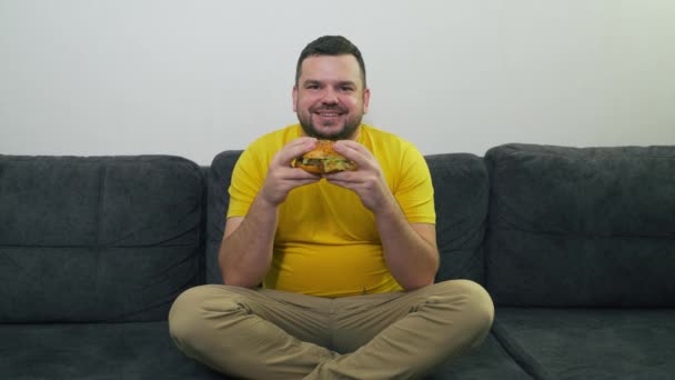 Pulchny młody człowiek z brązowymi oczami siedzi na szarej kanapie i je wielkiego tłustego hamburgera. Gryzie bułkę w duże kawałki. Pragnienie i głód. Cholesterol, niezdrowa dieta, niezdrowe odżywianie, obżarstwo — Wideo stockowe