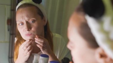 Genç kadın banyo aynasında kendine bakar, yüzünün derisini inceler, sivilceyi bulur ve sıkar. Yüzünde kızarıklık ve sivilceler var. Sağlık, kozmetoloji, cilt tedavisi