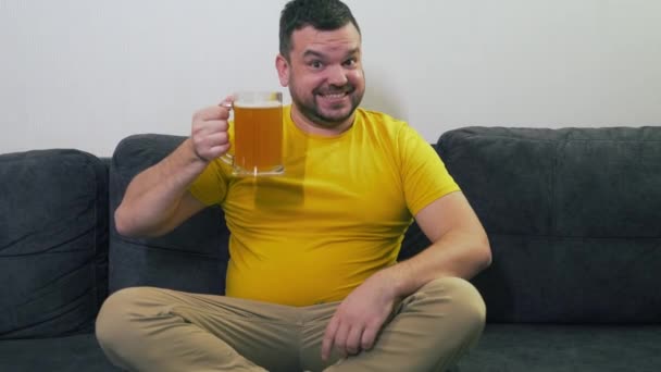 Молодий чоловік сидить на сірому дивані вдома перед телевізором, радіючи і п'ючи пиво зі скляного горнятка. Він п'є пиво у великих ярах і посміхається у всьому. Розслабся вдома, дивись телевізор. алкоголь — стокове відео