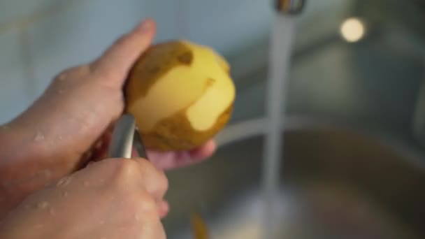 La donna sbuccia patate mature con pelapatate sopra il lavandino d'argento vicino all'acqua che scorre in cucina. Il dispositivo in metallo rimuove il sottile strato di patate. Note di alimentazione, verdure, cucina casalinga — Video Stock